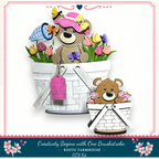 A wooden basket with a teddy bear and a small brown bear sitting inside, surrounded by flowers.