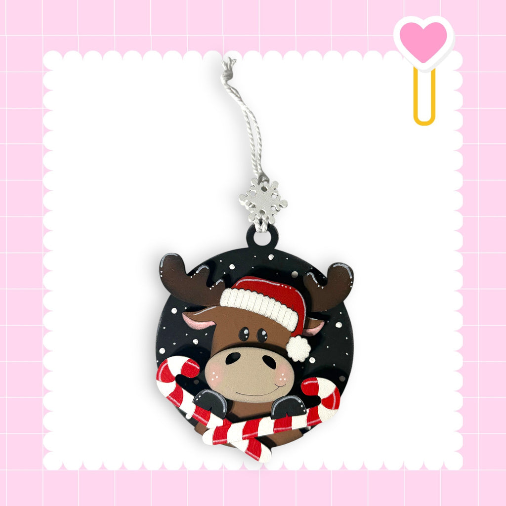 A Christmas ornament featuring a moose wearing a Santa hat and holding candy canes, set against a pink background with a scalloped edge.