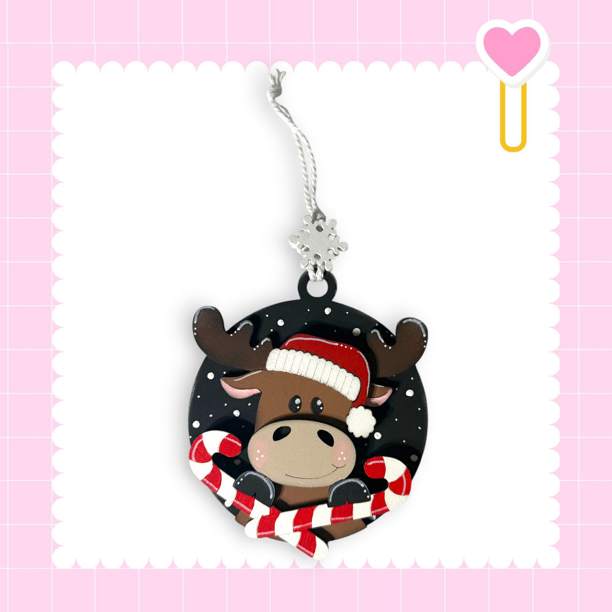 A Christmas ornament featuring a moose wearing a Santa hat and holding candy canes, set against a pink background with a scalloped edge.