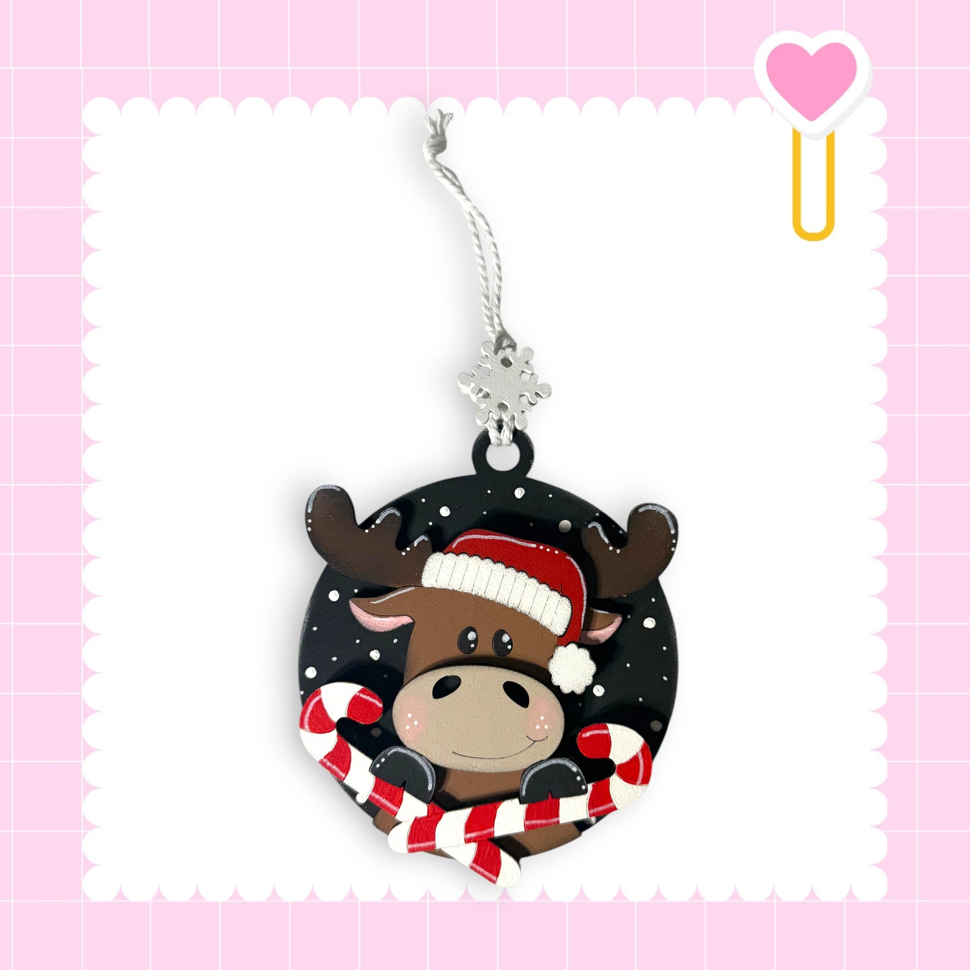 A Christmas ornament featuring a moose wearing a Santa hat and holding candy canes, set against a pink background with a scalloped edge.
