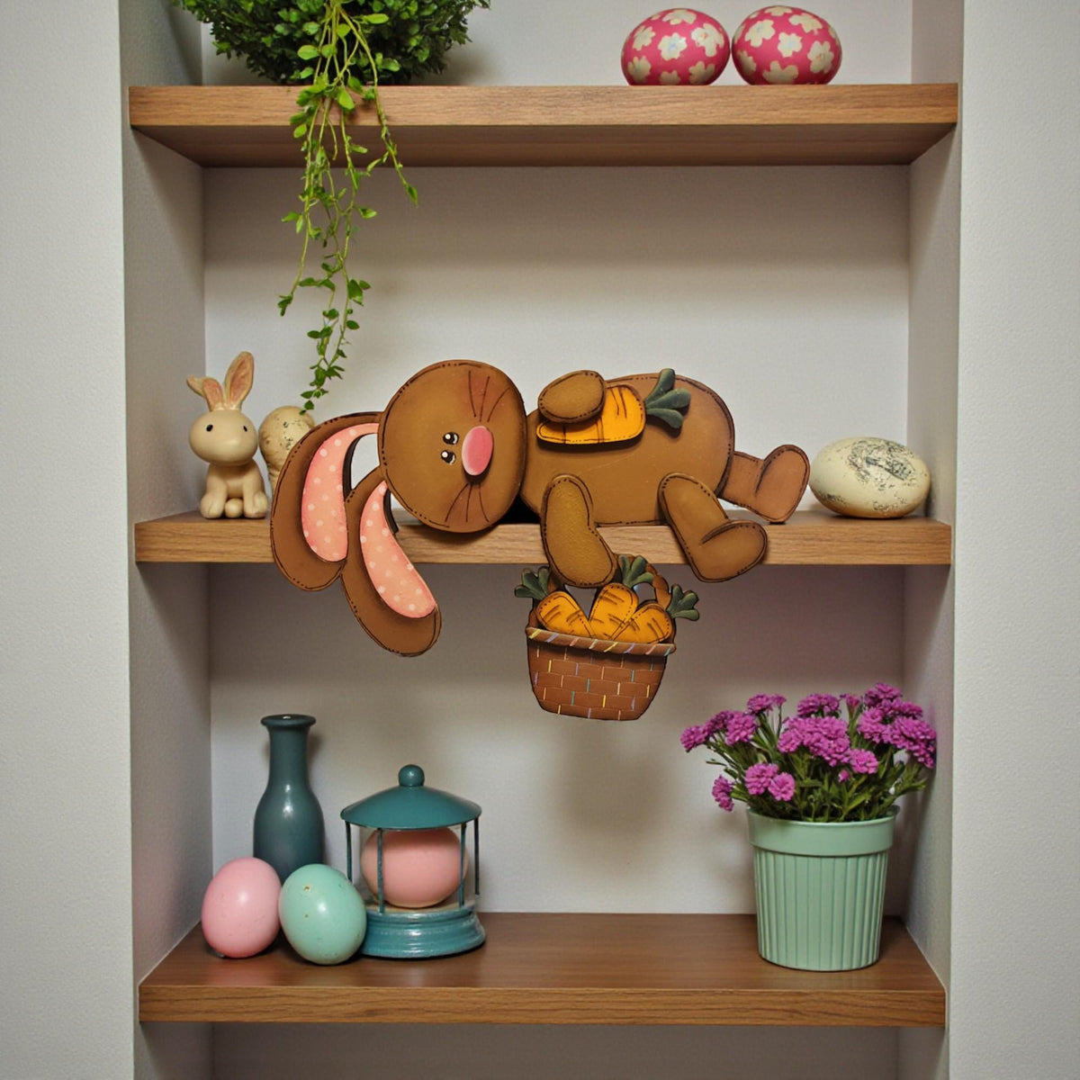 A wooden shelf displaying various Easter-themed items, including a hanging bunny figure, a basket of carrots, and a potted plant with purple flowers.
