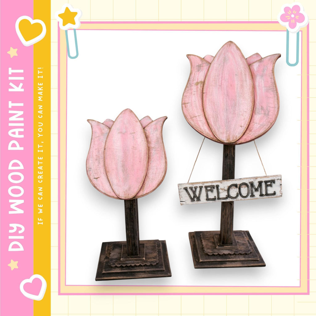 two wooden flower-shaped sculptures, each with a wooden sign attached to them. The sign reads "WELCOME". The sculptures are placed on a wooden base, and the background is a light pink color with a yellow border.