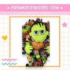 a Halloween-themed decoration with a green monster-like character holding a glittery pumpkin. The decoration is surrounded by a colorful border and includes a pink star and a heart.