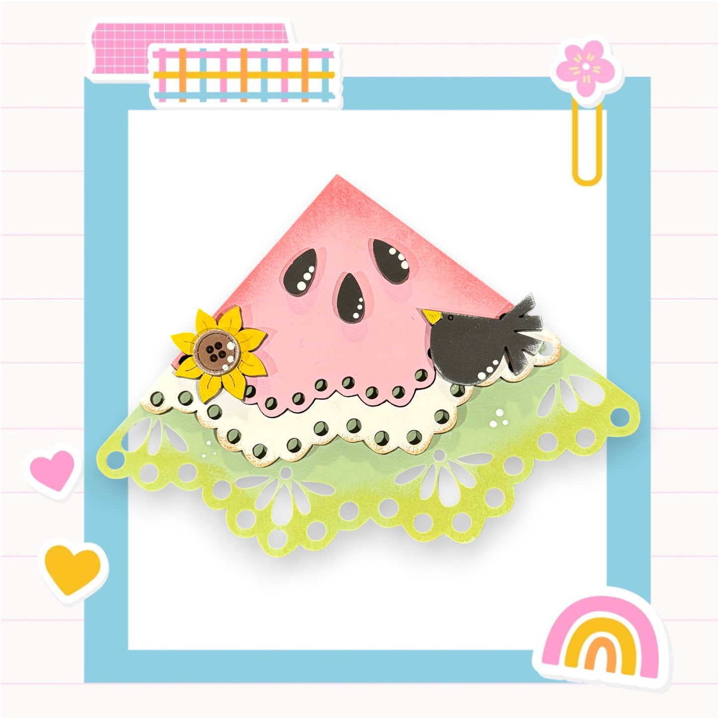A pink watermelon-shaped paper craft with a black bird perched on top, surrounded by various colorful and decorative elements such as a flower, a rainbow, and hearts.
