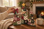 A hand is holding a red bow on a Christmas tree decoration, surrounded by other festive decorations on a table in front of a fireplace.