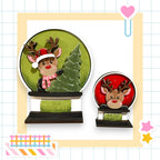 two deer figurines, one larger and one smaller, both wearing Santa hats and standing on a green snow globe. The larger deer is positioned on the left side of the snow globe, while the smaller deer is on the right side. The snow globe is surrounded by a yellow border, and there is a heart-shaped decoration on the right side of the image.