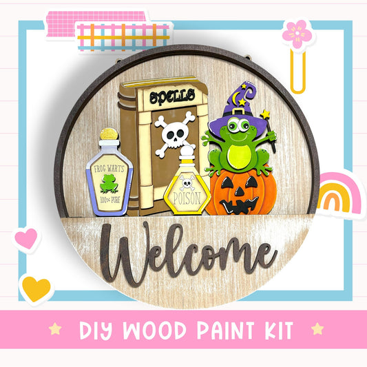 The image is a DIY wood paint kit featuring a welcome sign with a frog, a bottle of frog warts, a bottle of poison, a pumpkin, and a witch hat.