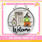 a DIY wood paint kit for creating a welcome sign. The kit includes a wooden sign with a cartoon frog, a skull and crossbones, a bottle of frog juice, and a pumpkin. The kit also includes the text "Welcome" and "DIY WOOD PAINT KIT" along with a rainbow and stars.