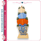 A hand-made wooden pumpkin display with three stacked pumpkins in a decorative vase, featuring a blue bow around the vase.