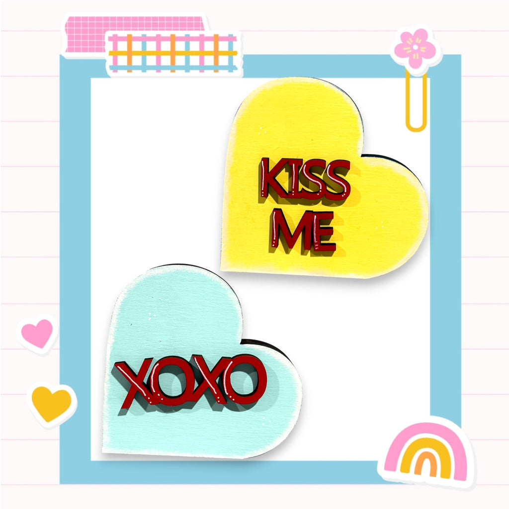 two heart-shaped cutouts with the text "KISS ME" and "XOXO" written on them, along with a pink flower and a rainbow-colored sticker.
