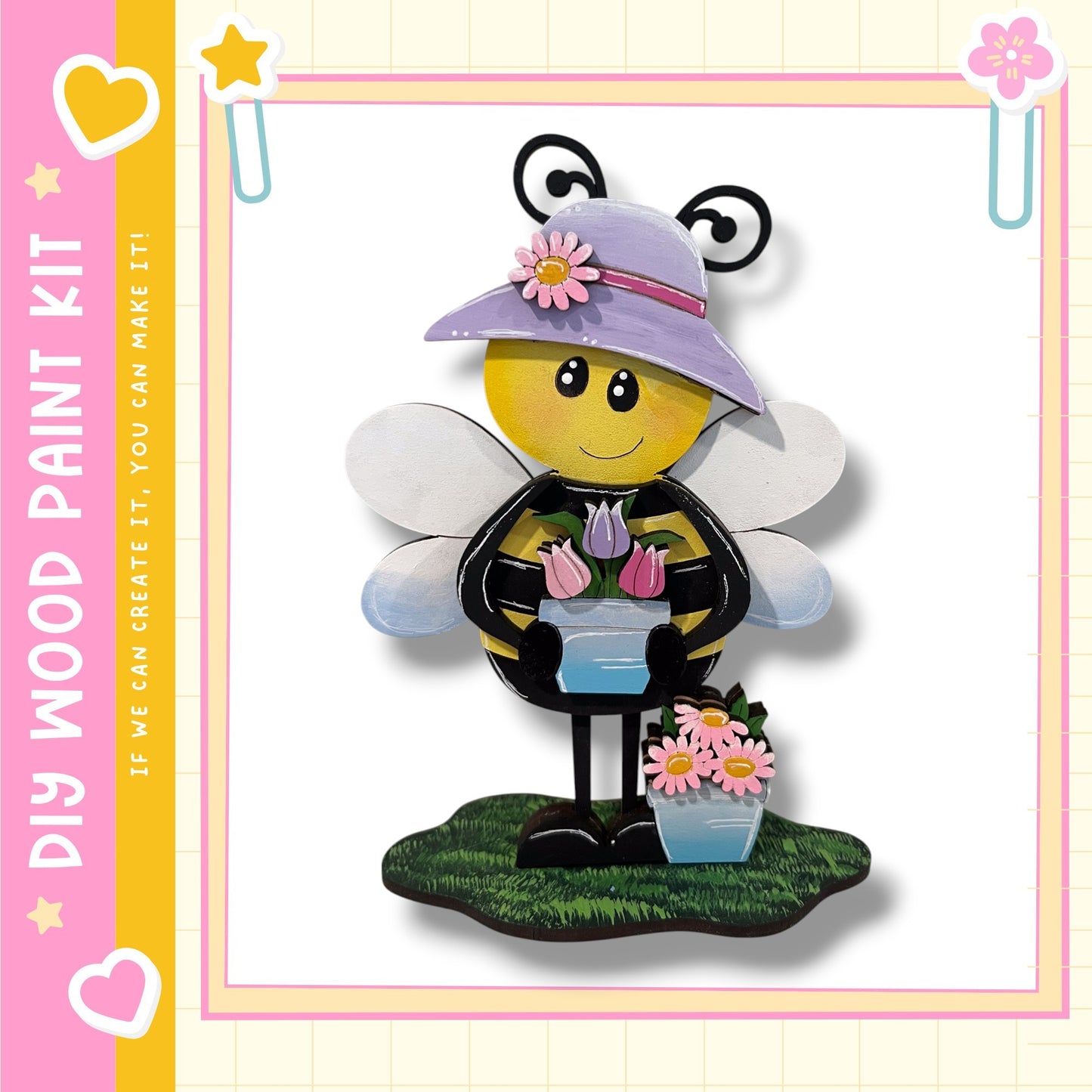 A wooden paint kit depicting a smiling bee wearing a purple hat and holding flowers, standing on a grassy surface.