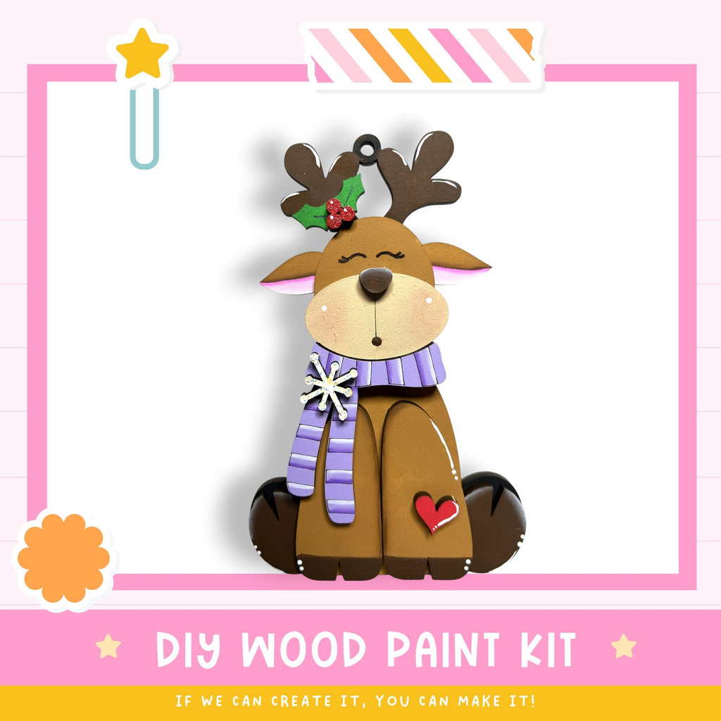 a wooden ornament in the shape of a reindeer, with a star decoration on its head and a heart-shaped ornament on its chest. The ornament is sitting on a pink background with a white border.
