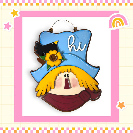 A colorful and whimsical scarecrow-like figure with a blue hat, yellow scarf, and a bird perched on top, set against a pink and yellow checkered background with stars and a rainbow.