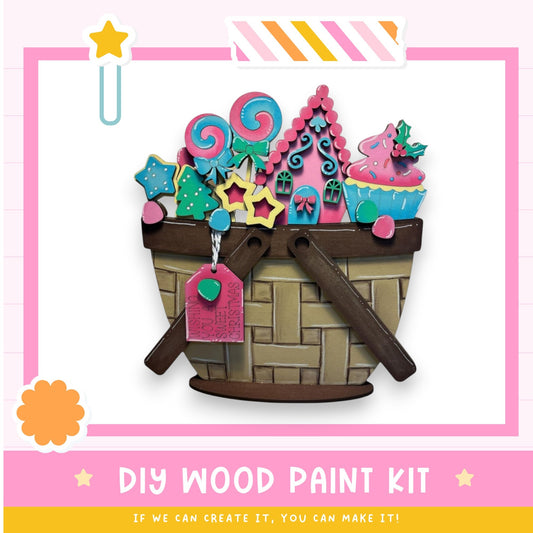 The image is a colorful and whimsical DIY wood paint kit, featuring a basket filled with various candy and cookie shapes, including lollipops, stars, and other festive decorations. The kit appears to be designed for children, as it is intended to be a fun and creative activity for them to engage in.