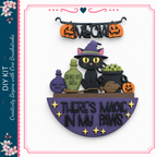 The image is a Halloween-themed DIY kit featuring a black cat wearing a witch's hat, surrounded by Halloween-themed objects such as pumpkins, a broomstick, and a cauldron. The kit includes a sign that says "There's Magic in My Paws" and a black cat figurine.