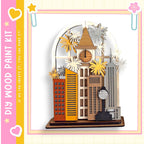 A wooden paint kit featuring a miniature city scene with various buildings, a clock, and decorative elements such as flowers and stars.