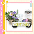 a wooden wagon filled with various Easter-themed decorations, including a bunny, flowers, and eggs. The wagon is labeled "HOPPY DAYS" and is surrounded by a pink and yellow border.
