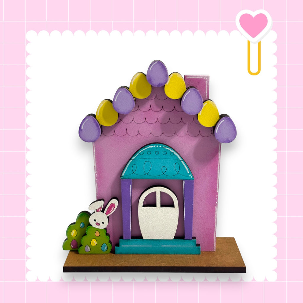 A colorful Easter-themed house with a bunny decoration, set against a pink background with a heart symbol.