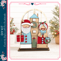 A wooden Christmas tree ornament featuring Santa Claus and an elderly woman, with a wreath and gift box. The ornament is displayed on a wooden base, and the background includes a blurred Christmas tree and other holiday decorations.