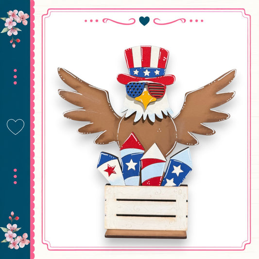 A wooden eagle wearing a patriotic hat and sunglasses, with its wings spread wide, is perched atop a wooden crate filled with fireworks.