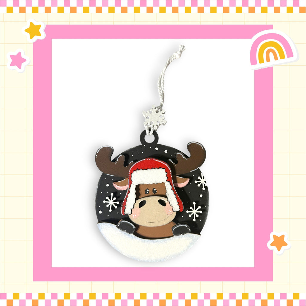 A decorative ornament featuring a cartoon moose wearing a red hat and surrounded by snowflakes, set against a pink and yellow checkered background with stars and a rainbow.
