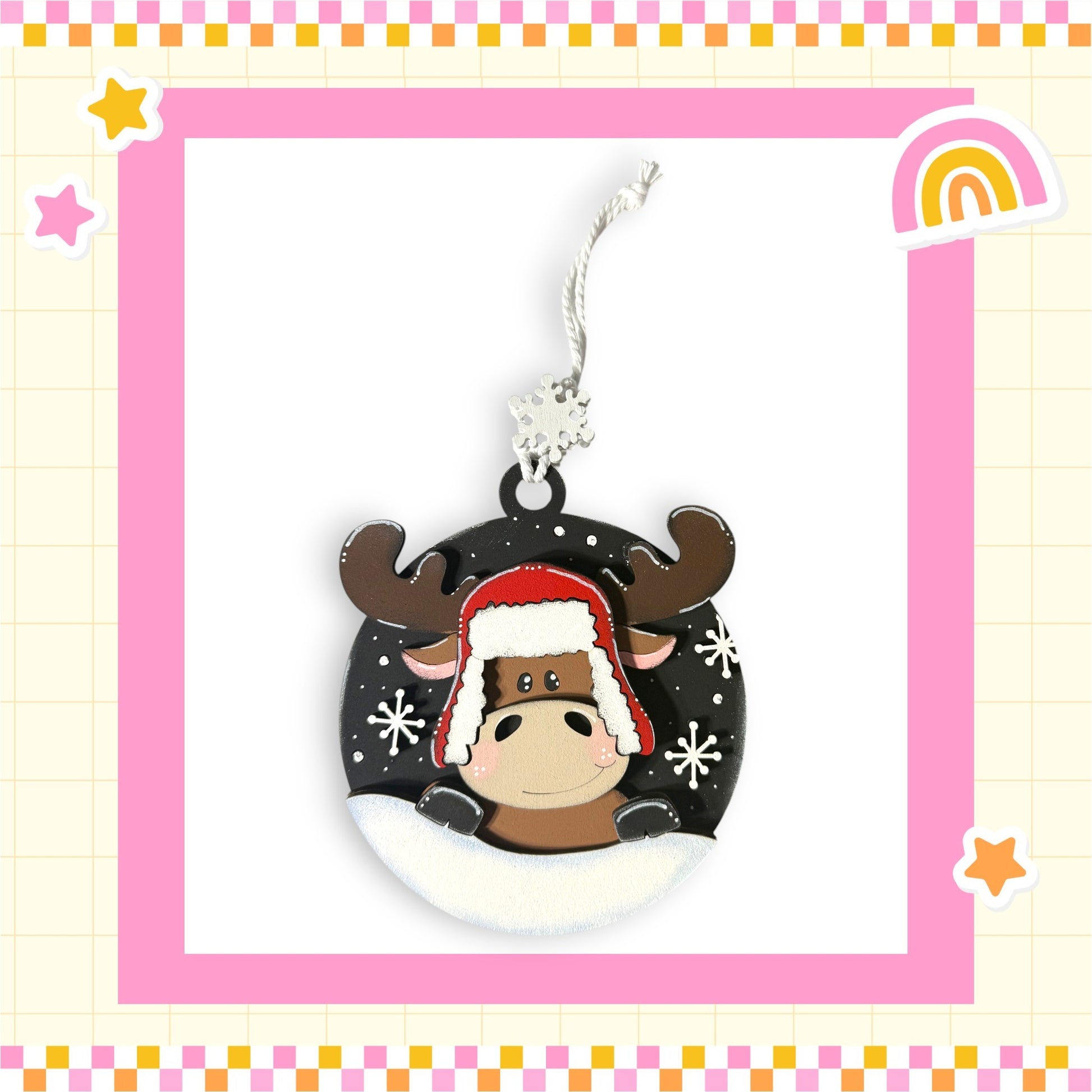 A decorative ornament featuring a cartoon moose wearing a red hat and surrounded by snowflakes, set against a pink and yellow checkered background with stars and a rainbow.