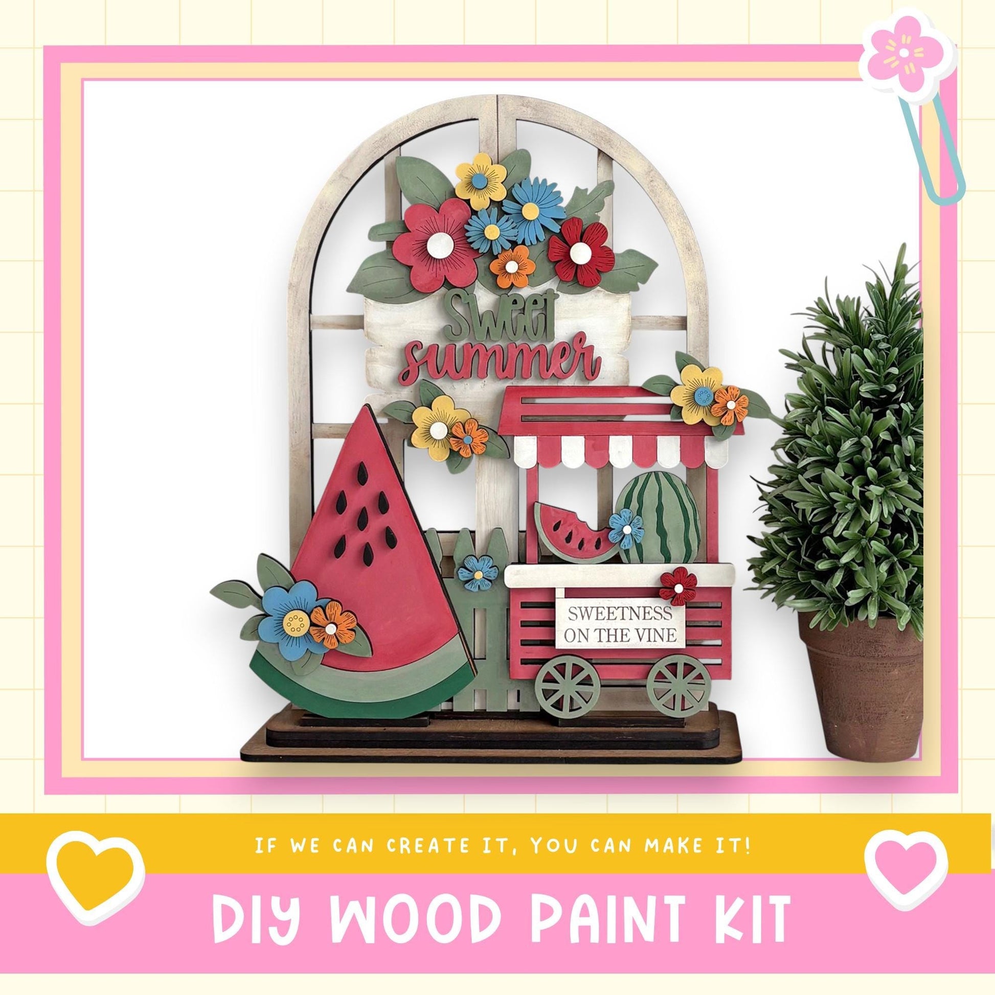 a DIY wood paint kit for creating a summer-themed decoration, including a wooden sign with the word "summer" and a wooden wagon with a watermelon on top. The kit is displayed in a pink frame, and there is a potted plant next to it.