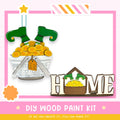 The image is a DIY wood paint kit for creating a wooden sign that says "HOME". The kit includes a wooden sign, a pair of green elf boots, and a basket filled with gold coins. The kit is designed to be a fun and creative project for anyone interested in woodworking and painting.