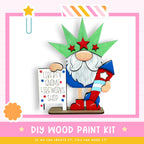 A wooden gnome figurine holding a sign that says "Liberty Gnome Fireworks Shop" and wearing a green hat with red stars. The gnome is standing on a pink and white striped base, and there is a star decoration above the gnome. The image also includes text that says "DIY WOOD PAINT KIT" and "IF WE CAN CREATE IT, YOU CAN MAKE IT!"