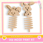 two wooden spring-like objects, resembling toy rabbits, with a pink and yellow border. The text below the image reads "DIY WOOD PAINT KIT" and "If we can create it, you can make it!"
