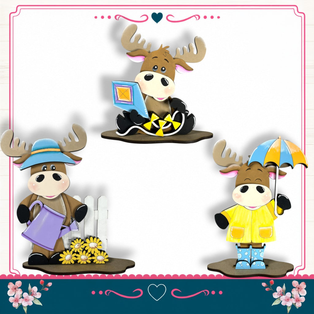 three moose figurines dressed in different outfits, each holding an umbrella and a flower, set against a white background with a pink border.