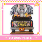 A wooden toy truck decorated with a bat and various candy items, including a sign that says "BABY FOR CANDY". The truck is set against a pink and yellow background with stars and a rainbow.