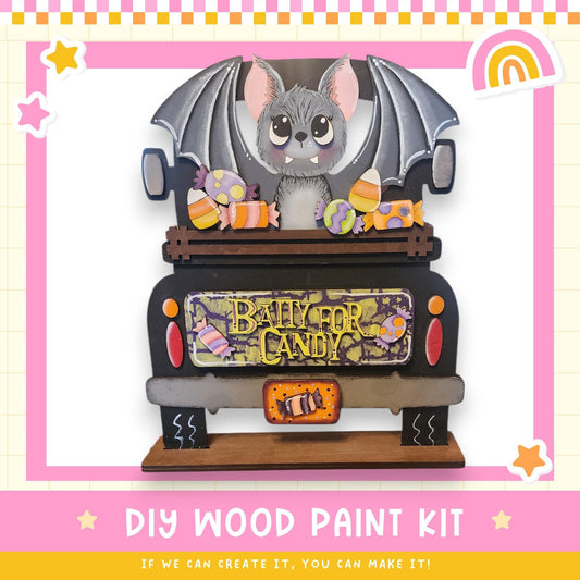 A wooden toy truck decorated with a bat and various candy items, including a sign that says "BABY FOR CANDY". The truck is set against a pink and yellow background with stars and a rainbow.