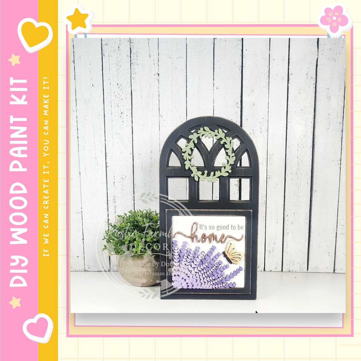 a black wooden frame with a wreath hanging on it, placed on a white shelf against a white wooden wall. Next to the frame is a potted plant with purple flowers. The frame has a sign with the text "It's good to be home" written on it.