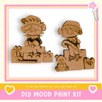 two wooden figures, likely representing a couple, sitting on a wooden base. The figures are decorated with bows and other embellishments, suggesting they are intended as a gift or decoration. The image also includes text that reads "DIY WOOD PAINT KIT" and "If we can create it, you can make it!" This indicates that the image is likely a promotional material for a DIY wood paint kit, encouraging viewers to create their own wooden figures.