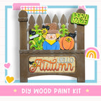 The image is a DIY wood paint kit for creating a fall-themed decoration, featuring a scarecrow, corn maze, and a sign that says "Hello Autumn".