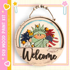 A wooden sign featuring a cartoon depiction of the Statue of Liberty surrounded by various American symbols, including fireworks and a banner that says "Welcome".