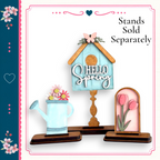 a wooden birdhouse with a "Hello Spring" sign on top, a blue watering can with flowers, and a pink flower arrangement. The birdhouse and watering can are placed on a wooden stand, and the pink flower arrangement is displayed in a frame.
