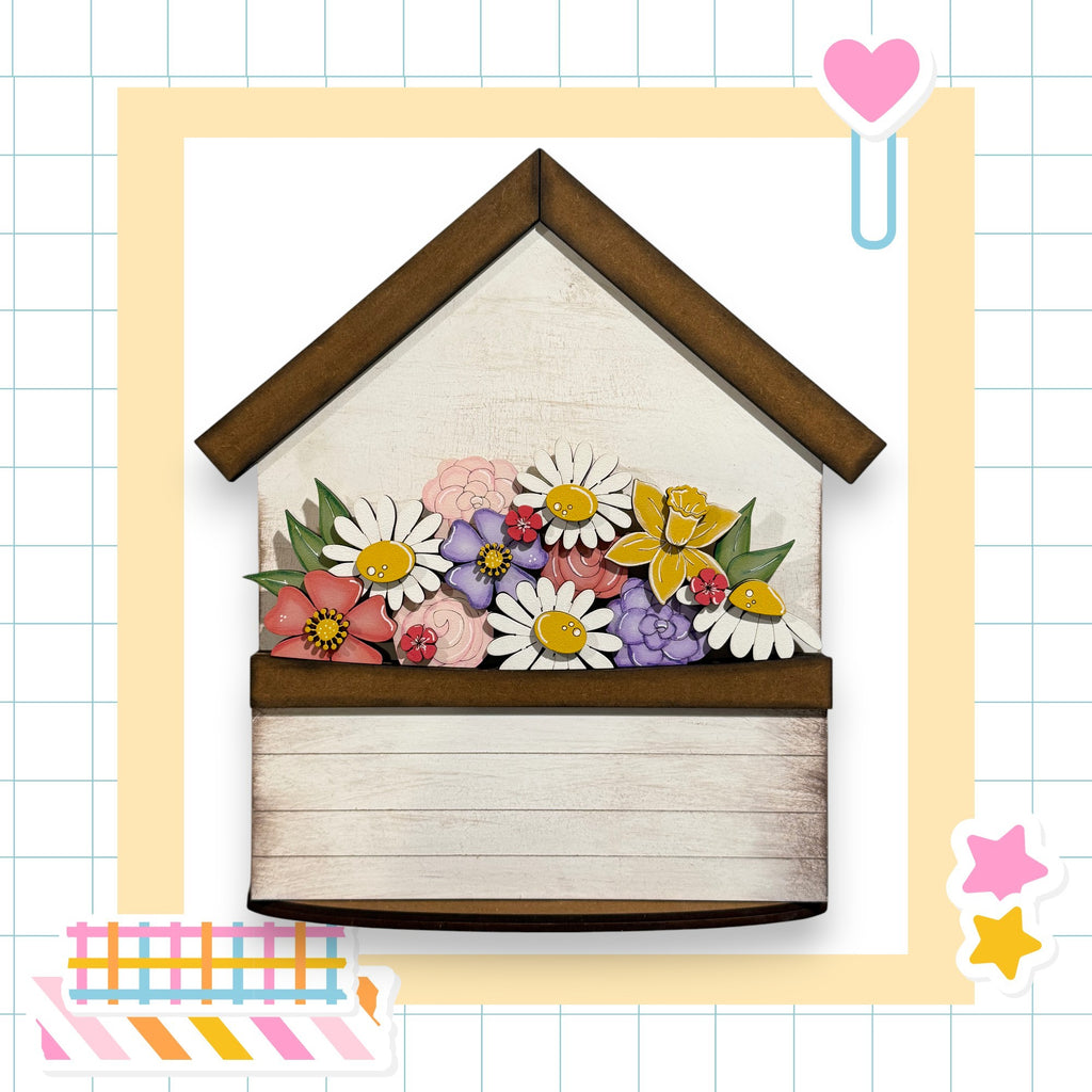 A wooden house-shaped planter filled with a variety of colorful flowers, including daisies, roses, and other blooms.
