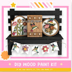 The image is a DIY wood paint kit featuring a wooden bench with various decorative elements, including a wooden sign with a bird and flowers, hanging banners with colorful flowers, and a wooden box with a bird and flowers.