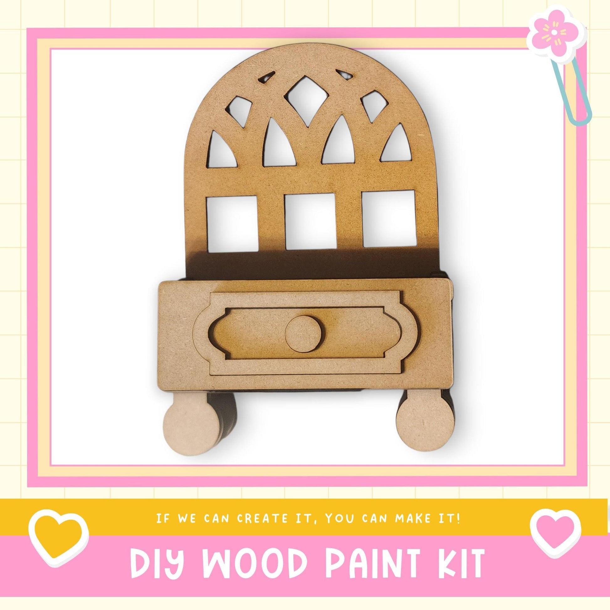 a DIY wood paint kit, with a wooden cabinet or shelf depicted in the center. The kit appears to be made of wood and has a drawer. The image is framed with a pink border and includes a heart symbol on the left side. The text "DIY WOOD PAINT KIT" is displayed at the bottom of the image.