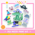 The image is a colorful and playful DIY wood paint kit featuring a green alligator wearing various hats and accessories, including a pirate hat, a top hat, and a cat's hat.