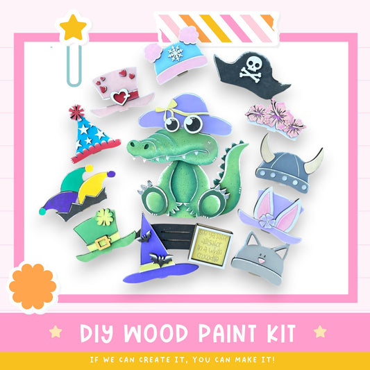 The image is a colorful and playful DIY wood paint kit featuring a green alligator wearing various hats and accessories, including a pirate hat, a top hat, and a cat's hat.