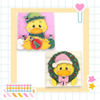 two cheerful cartoon duck characters, one wearing a Santa hat and the other a wreath, set against a colorful background.