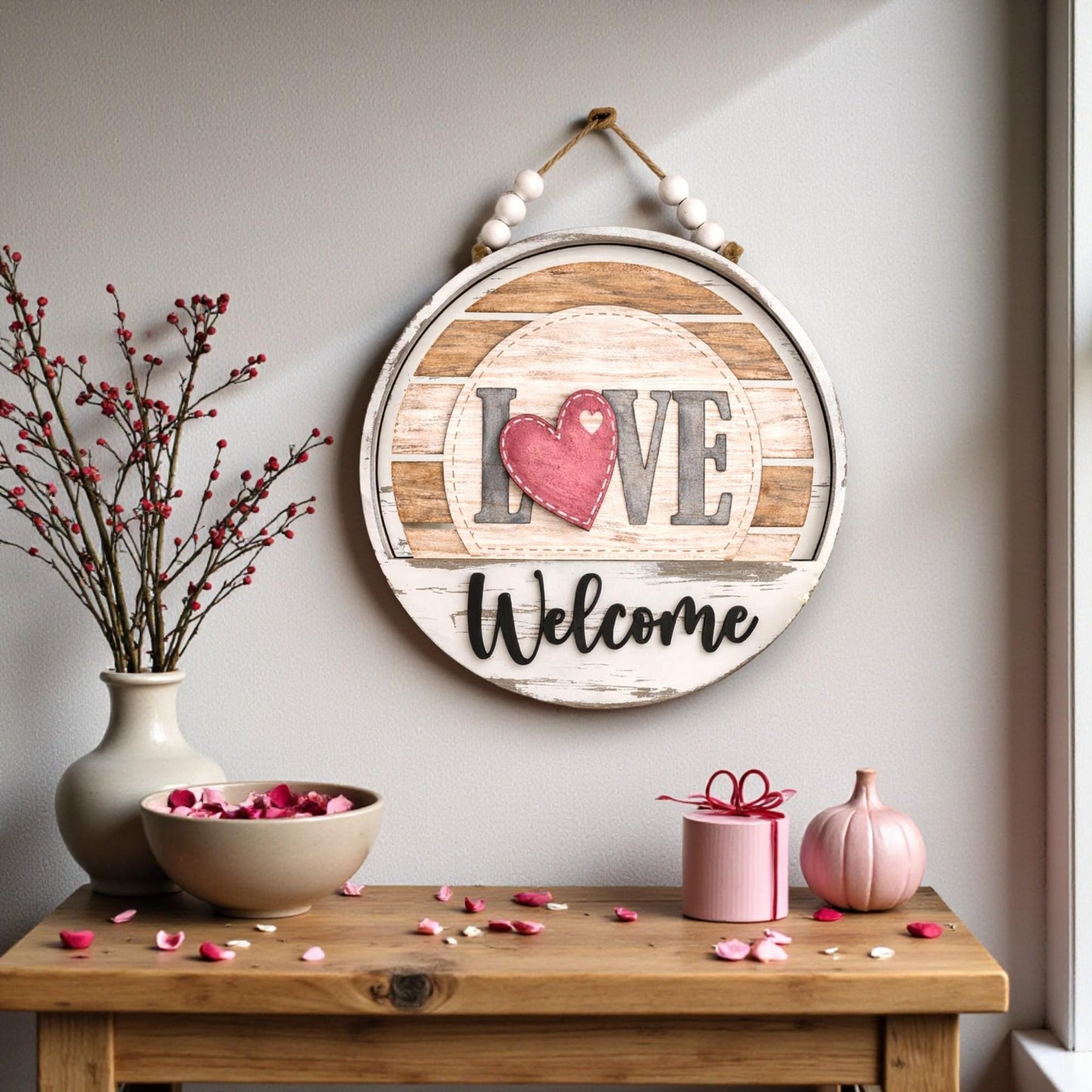 a wooden table with a bowl of pink flowers, a vase, and a small pink pumpkin. Above the table, there is a wooden sign with the word "Love" and the word "Welcome" written on it.