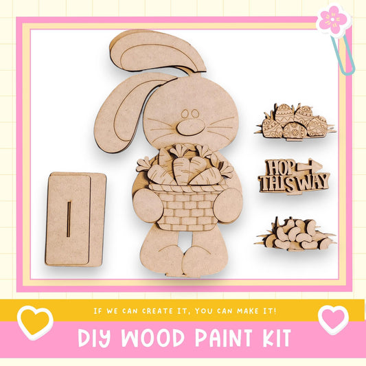 The image is a DIY wood paint kit featuring a wooden rabbit holding a basket of carrots, along with various other wooden shapes and a sign that says "HOP THIS WAY".