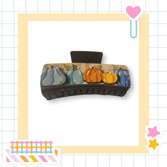 a black hair clip with a pumpkin and blue squash design on it, set against a white background with a grid pattern.