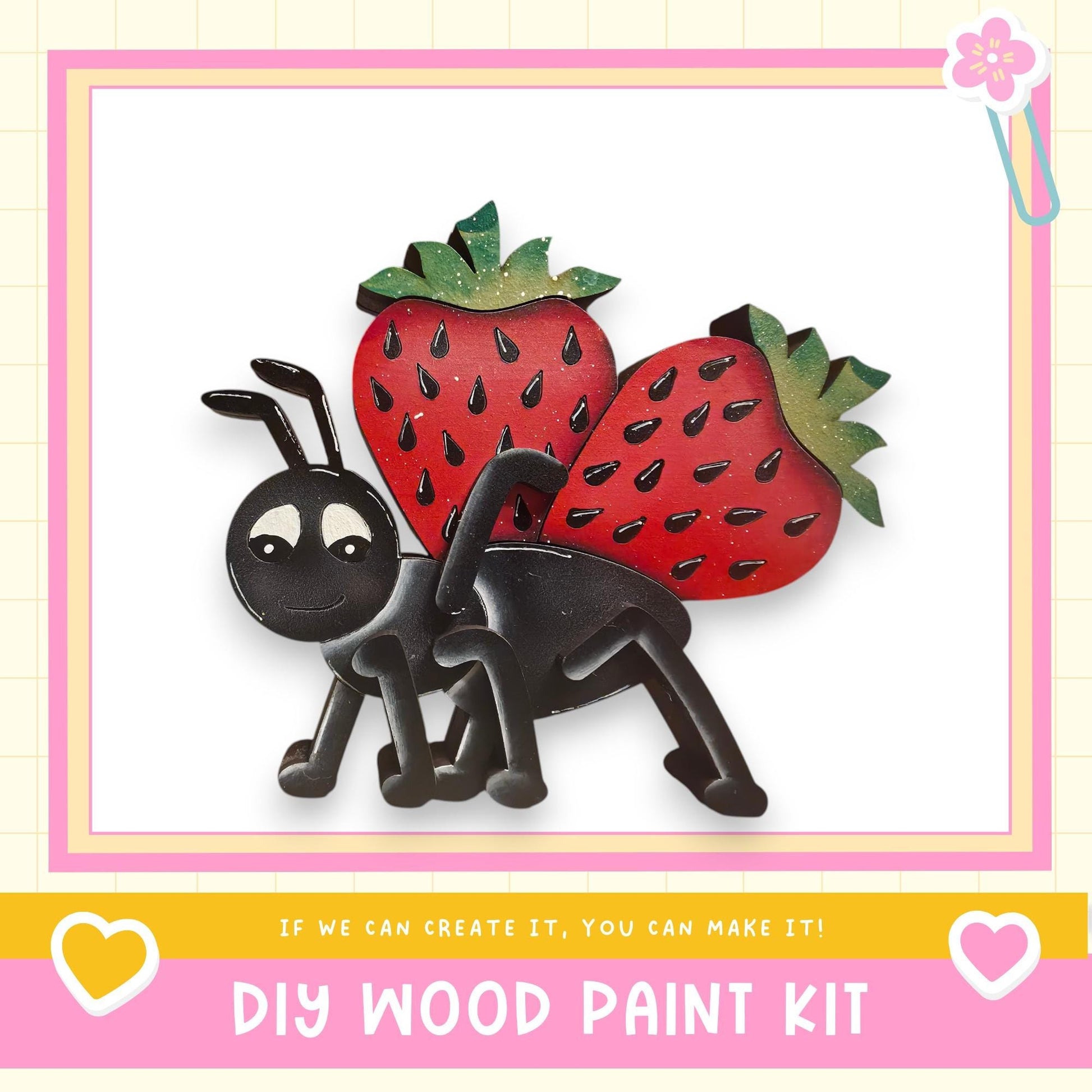 The image is a DIY wood paint kit featuring a black ant with red strawberries on its back, set against a pink and yellow background.