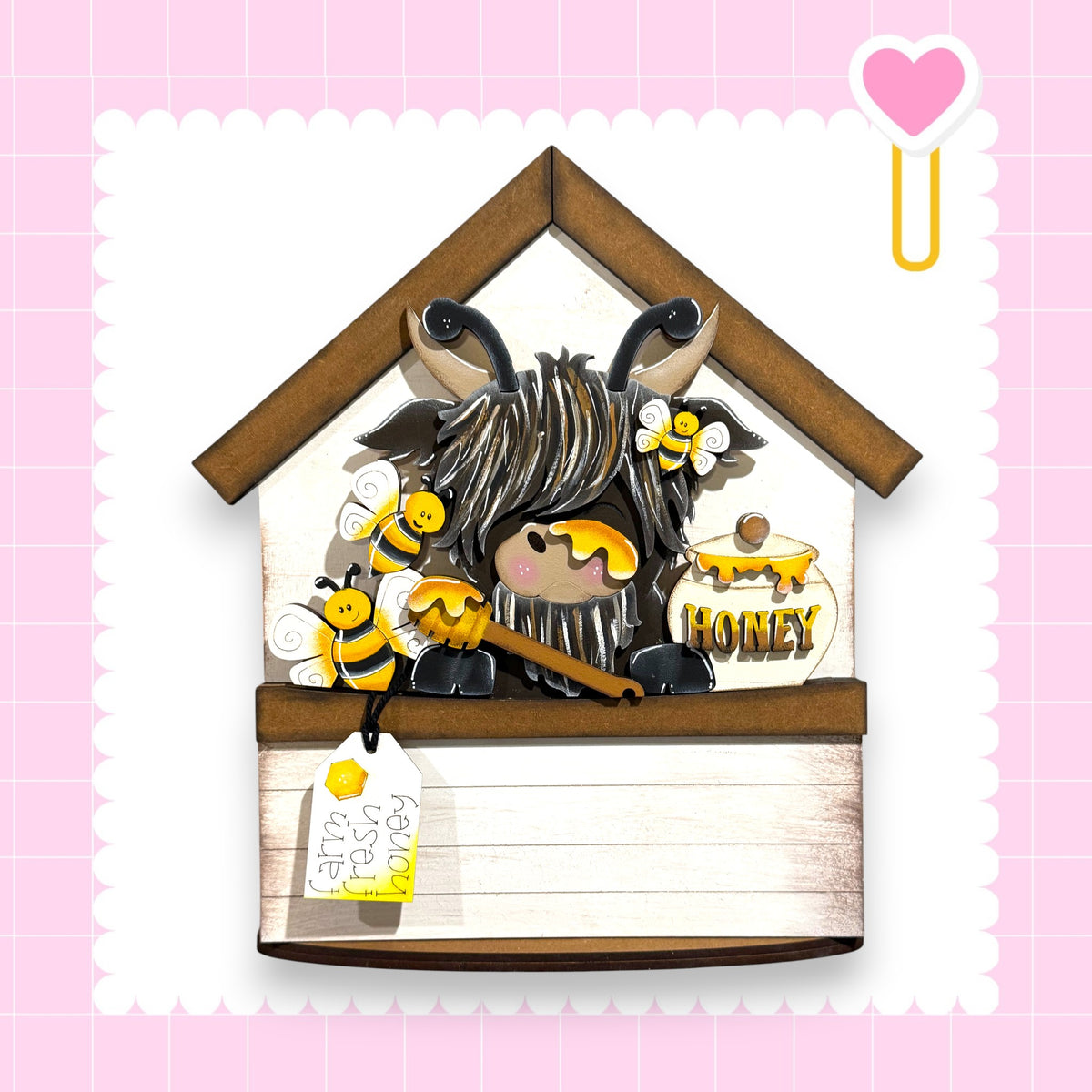 A wooden house-shaped object with a honey jar, a bee, and a tag attached to it, set against a pink background with a heart symbol.