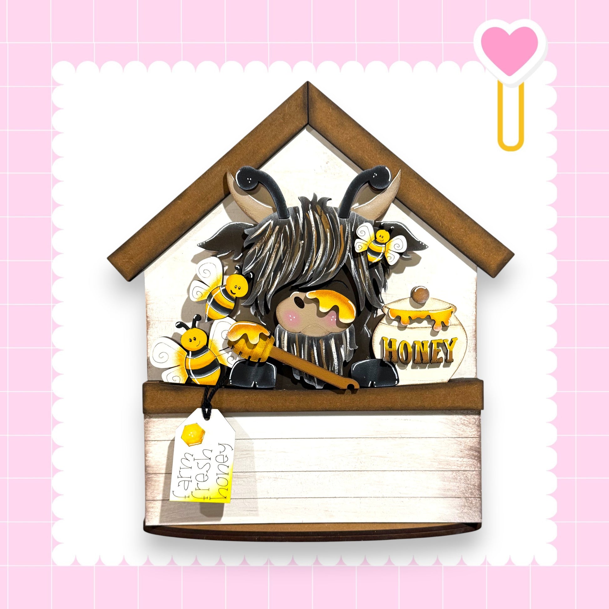 A wooden house-shaped object with a honey jar, a bee, and a tag attached to it, set against a pink background with a heart symbol.